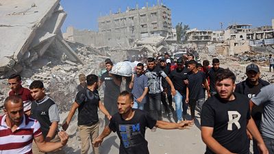 Hombres en duelo transportan un cuerpo envuelto en tela blanca por una calle llena de escombros y edificios destruidos. Se observa una 'R' blanca en una camiseta y la palabra "WIND" en otra. Hombres en duelo transportan un cuerpo envuelto en tela blanca por una calle llena de escombros y edificios destruidos. Se observa una 'R' blanca en una camiseta y la palabra "WIND" en otra.