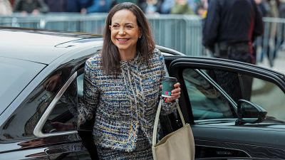 María Corina Machado, con una sonrisa, sale de un coche, vestida con un conjunto de tweed y un bolso beige, portando un vaso de café.