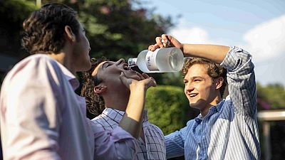 Jóvenes disfrutando en un entorno exterior; uno vierte líquido de una botella a otro, mientras un tercero observa. Escena festiva con implicaciones negativas. Jóvenes disfrutando en un entorno exterior; uno vierte líquido de una botella a otro, mientras un tercero observa. Escena festiva con implicaciones negativas.