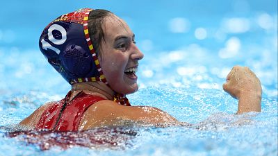 En la imagen, una jugadora de waterpolo, con el número 10 en su gorro, celebra un gol en el agua, sonriendo y levantando el puño. El gorro es azul oscuro con detalles y el bañador es rojo.