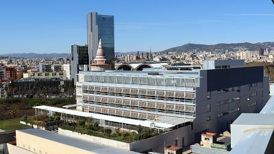 Una vista panorámica de Barcelona muestra el Hospital del Mar en primer plano, un edificio moderno con numerosas ventanas y una sección gris oscura, con el horizonte de la ciudad y la Sagrada Familia al fondo. Una vista panorámica de Barcelona muestra el Hospital del Mar en primer plano, un edificio moderno con numerosas ventanas y una sección gris oscura, con el horizonte de la ciudad y la Sagrada Familia al fondo.