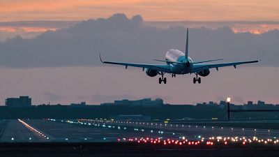 Un avión de pasajeros, con detalles en rojo y blanco, se aproxima a la pista de aterrizaje iluminada, con un cielo teñido de tonos rosados y anaranjados, posiblemente al amanecer o atardecer, y un horizonte urbano al fondo. Un avión de pasajeros, con detalles en rojo y blanco, se aproxima a la pista de aterrizaje iluminada, con un cielo teñido de tonos rosados y anaranjados, posiblemente al amanecer o atardecer, y un horizonte urbano al fondo.