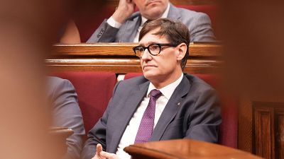 Primer pla de Salvador Illa, polític espanyol, assegut en un hemicicle. Porta ulleres, vestit fosc i corbata morada. Entorn parlamentari amb seients vermells i fusta fosca al fons. Primer pla de Salvador Illa, polític espanyol, assegut en un hemicicle. Porta ulleres, vestit fosc i corbata morada. Entorn parlamentari amb seients vermells i fusta fosca al fons.