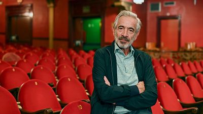 En un teatro con butacas rojas vacías, un hombre con camisa azul claro y chaqueta oscura posa con los brazos cruzados, posiblemente en un momento de descanso o espera.