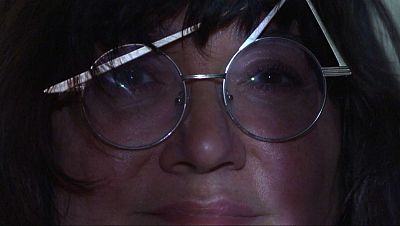 Primer plano del rostro de una mujer con gafas redondas y montura metálica, posiblemente una cineasta, con una patilla de las gafas levantada, capturado con una iluminación tenue que genera sombras.