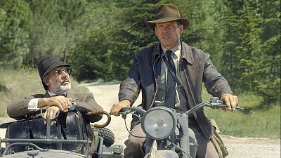 Indiana Jones (Harrison Ford) en motocicleta con sidecar, donde viaja acompañado por un hombre con traje y bastón (Sean Connery). Escena en camino de tierra arbolado.