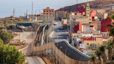 Una carretera paralela a una valla metálica fronteriza, con vegetación baja a los lados y edificios de varios colores al fondo, bajo un cielo despejado.