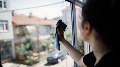 Una mujer joven limpia desde dentro el cristal de una ventana, en la que se ve la calle de un barrio periférico residencial. Una mujer joven limpia desde dentro el cristal de una ventana, en la que se ve la calle de un barrio periférico residencial.