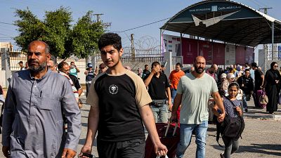 Un grupo de personas caminan al aire libre, con un hombre barbudo y un joven con maleta en primer plano. Al fondo, se distingue un edificio con un letrero. Un grupo de personas caminan al aire libre, con un hombre barbudo y un joven con maleta en primer plano. Al fondo, se distingue un edificio con un letrero.