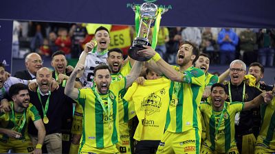 Un grupo de jugadores de fútbol sala, vestidos con camisetas amarillas y verdes, celebran la victoria levantando una copa plateada, con un ambiente de euforia y alegría.