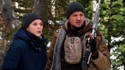 Elizabeth Olsen y Jeremy Renner, en escena de *Wind River*,  en un bosque nevado; ambos con abrigos y gorros, muestran preocupación y tensión.