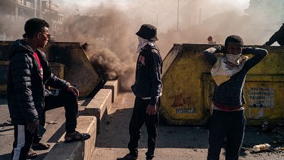 En una escena urbana con humo, tres jóvenes se encuentran frente a contenedores con grafitis. Uno lleva gafas de sol y chaqueta oscura, otro un sombrero y sudadera, y el tercero una camiseta clara, todos con el rostro parcialmente cubierto.