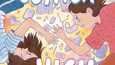 En la portada de un cómic, dos chicas jóvenes se relajan sobre un fondo amarillo claro, rodeadas de objetos como cuadernos, un teléfono móvil y una mochila, sugiriendo un ambiente escolar y juvenil.