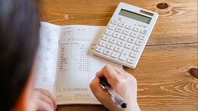 En la imagen, una persona escribe en un libro abierto que parece ser un registro de gastos, con números y texto en japonés, junto a una calculadora.