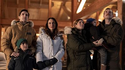 Un grupo familiar, compuesto por dos parejas y dos niños, observa algo fuera de la imagen. Los adultos visten chaquetas de invierno y los niños gorros, en un entorno con techo de madera, sugiriendo un ambiente frío y familiar. Un grupo familiar, compuesto por dos parejas y dos niños, observa algo fuera de la imagen. Los adultos visten chaquetas de invierno y los niños gorros, en un entorno con techo de madera, sugiriendo un ambiente frío y familiar.