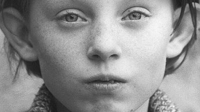 Primer plano en blanco y negro del rostro de un niño o niña, con ojos claros y labios finos, mirando directamente a la cámara. La imagen tiene una textura granulada. Primer plano en blanco y negro del rostro de un niño o niña, con ojos claros y labios finos, mirando directamente a la cámara. La imagen tiene una textura granulada.