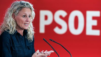 Montse Mínguez, con camisa vaquera, habla ante micrófonos en una rueda de prensa. Se observa el logo de un partido político detrás de ella.