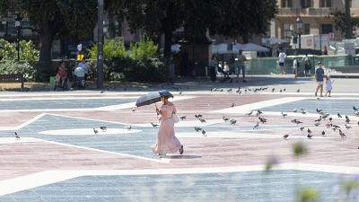 Mujer con paraguas oscuro se protege del sol en una plaza con suelo geométrico y palomas. Al fondo, árboles, bancos y edificios urbanos. Mujer con paraguas oscuro se protege del sol en una plaza con suelo geométrico y palomas. Al fondo, árboles, bancos y edificios urbanos.
