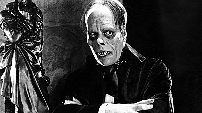 En la fotografía en blanco y negro, un hombre con maquillaje pálido y deformado, con los brazos cruzados, personifica a un personaje con ojos hundidos y dentadura visible, vestido con traje oscuro y corbata negra, en un ambiente sombrío.