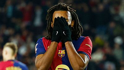 Futbolista con camiseta azulgrana y guantes negros se cubre el rostro mostrando decepción. Otro jugador se ve al fondo. Futbolista con camiseta azulgrana y guantes negros se cubre el rostro mostrando decepción. Otro jugador se ve al fondo.