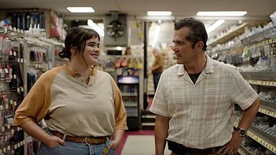 Escena en una ferretería: Benicio del Toro conversa con una mujer joven con camisa beige y pantalones vaqueros. Fondo con estanterías y gente. Escena en una ferretería: Benicio del Toro conversa con una mujer joven con camisa beige y pantalones vaqueros. Fondo con estanterías y gente.