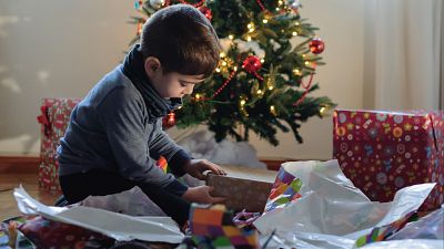 Niño pequeño abriendo regalos junto a un árbol de Navidad decorado, rodeado de papel de regalo de colores.  Escena llena de ilusión.