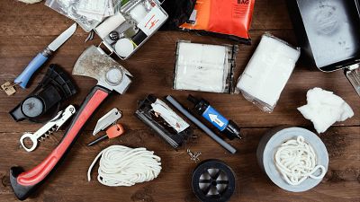 Variedad de herramientas y suministros para supervivencia a 72 horas: hacha, navaja, pedernal, pastillas, filtro de agua, cinta, cuerda y más, dispuestos sobre madera.