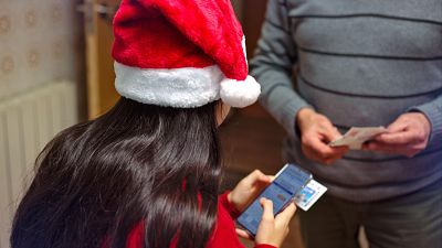 Una persona con gorro navideño compra Lotería de Navidad online mientras otra sujeta décimos físicos.