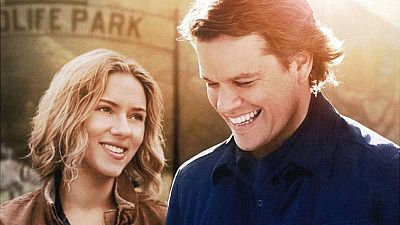 Scarlett Johansson, con chaqueta marrón, y Matt Damon, con camisa azul, sonríen mutuamente en una escena con un cartel desgastado que dice "OLIFE PARK" al fondo. Scarlett Johansson, con chaqueta marrón, y Matt Damon, con camisa azul, sonríen mutuamente en una escena con un cartel desgastado que dice "OLIFE PARK" al fondo.