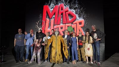 Grupo de personas e invitados caracterizados como criaturas de terror posan frente a un gran logotipo rojo de 'MAD HORROR' en un escenario.