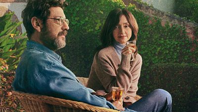 Javier Cámara y una mujer, con copas en mano, sentados en sillas de mimbre en un entorno tranquilo y verde. Javier Cámara y una mujer, con copas en mano, sentados en sillas de mimbre en un entorno tranquilo y verde.