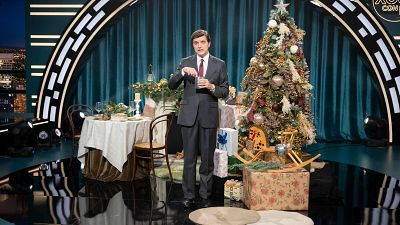 En un escenario decorado para la Navidad, un hombre vestido con traje y corbata sostiene un vaso. A su alrededor se observan elementos festivos como un árbol de Navidad y una mesa con adornos, con un fondo de cortinas y luces.