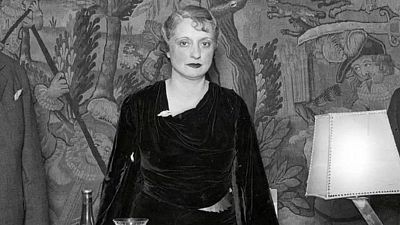 Retrato en blanco y negro de Mari Luz Morales con vestido oscuro, sentada en un evento social con una copa y botella en la mesa cercana. Fondo con tapiz y otras personas.