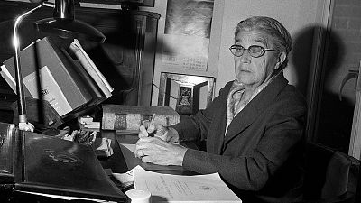 María Moliner, en blanco y negro, sentada en su despacho trabajando entre libros y papeles.  Imagen que refleja su dedicación a la lexicografía.