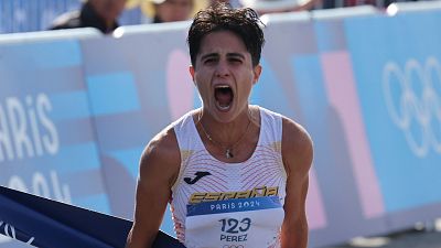Atleta cruzando la meta con expresión de triunfo; camiseta blanca con detalles rojos y dorados, dorsal 123; fondo con valla publicitaria de París 2024. Atleta cruzando la meta con expresión de triunfo; camiseta blanca con detalles rojos y dorados, dorsal 123; fondo con valla publicitaria de París 2024.