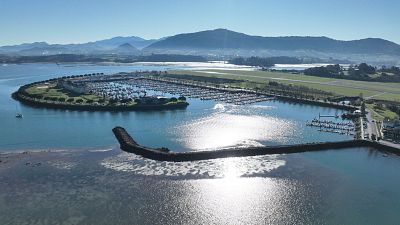 Una vista general de un puerto deportivo en una bahía de aguas tranquilas, con barcos atracados y montañas al fondo. El cielo es azul y despejado, y una escollera protege el puerto.