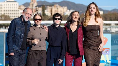 El equipo de la película "Yo no moriré de amor" posa para una foto en un festival de cine. Se observan cinco personas, con vestimentas variadas, frente a un cartel y un paisaje urbano con montañas. El equipo de la película "Yo no moriré de amor" posa para una foto en un festival de cine. Se observan cinco personas, con vestimentas variadas, frente a un cartel y un paisaje urbano con montañas.