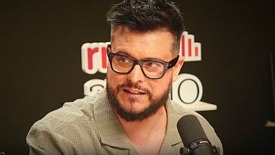 Martin Barreiro, presentador con gafas y barba, en un estudio de radio con logo y micrófono. Camisa verde oliva. Martin Barreiro, presentador con gafas y barba, en un estudio de radio con logo y micrófono. Camisa verde oliva.
