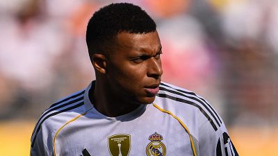 Futbolista con camiseta blanca del Real Madrid, escudo visible y parche dorado. Expresión seria, fondo desenfocado con tonos naranja, blanco y rosa. Futbolista con camiseta blanca del Real Madrid, escudo visible y parche dorado. Expresión seria, fondo desenfocado con tonos naranja, blanco y rosa.