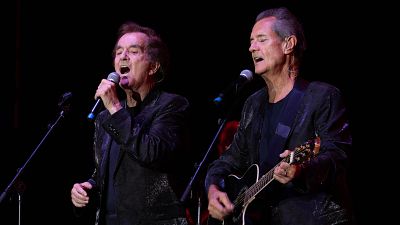 Concierto del Dúo Dinámico: Manuel de la Calva canta con micrófono en mano mientras Ramón Arcusa toca la guitarra electroacústica; ambos con chaquetas oscuras. Concierto del Dúo Dinámico: Manuel de la Calva canta con micrófono en mano mientras Ramón Arcusa toca la guitarra electroacústica; ambos con chaquetas oscuras.