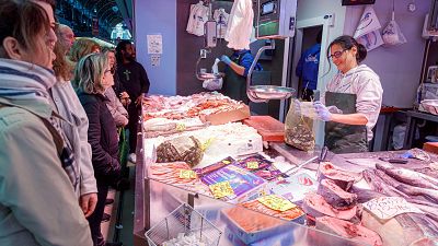 Una pescadería concurrida muestra a una empleada atendiendo a los clientes tras un mostrador repleto de pescado y marisco, como atún y salmón, con precios a la vista. Otro empleado se encuentra en la sección de carnes.