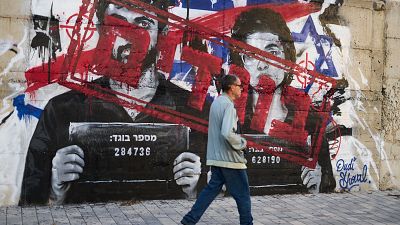 Grafiti en hormigón con retratos de Yuval Abraham y Rachel Szor, marcados con "X" rojas y símbolos de la bandera de Israel. Sostienen carteles con "מספר בוגד:" y números. Firma "Dudi Shoval" en azul.
