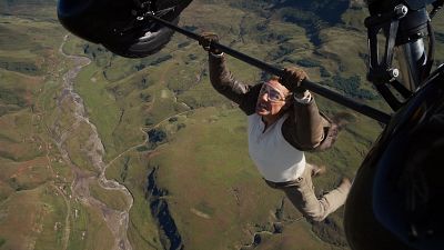 Actor colgado de una barra a gran altura, con gafas protectoras, camiseta blanca y pantalones beige, sobre un paisaje montañoso. Actor colgado de una barra a gran altura, con gafas protectoras, camiseta blanca y pantalones beige, sobre un paisaje montañoso.