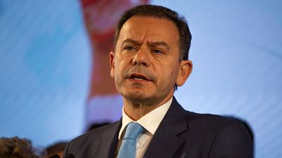 Ex primer ministro montenegrino, en traje oscuro y corbata azul, habla ante micrófonos en un evento con fondo azul claro. Ex primer ministro montenegrino, en traje oscuro y corbata azul, habla ante micrófonos en un evento con fondo azul claro.