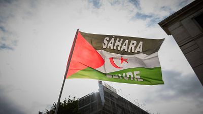 Bandera del Sáhara Occidental con "SÁHARA LIBRE" en blanco, triángulo rojo, franjas de colores, cielo nublado y siluetas urbanas. Bandera del Sáhara Occidental con "SÁHARA LIBRE" en blanco, triángulo rojo, franjas de colores, cielo nublado y siluetas urbanas.