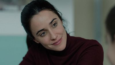Primer plano de una mujer con cabello oscuro recogido y una sonrisa suave, vestida con un jersey de cuello alto. El fondo difuminado muestra tonos claros y una pared azul pálido. Primer plano de una mujer con cabello oscuro recogido y una sonrisa suave, vestida con un jersey de cuello alto. El fondo difuminado muestra tonos claros y una pared azul pálido.