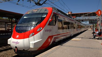 Tren de Renfe blanco y rojo en andén con hombre sentado, banco rojo y señal de andén 1. Puente peatonal naranja al fondo bajo cielo azul. Tren de Renfe blanco y rojo en andén con hombre sentado, banco rojo y señal de andén 1. Puente peatonal naranja al fondo bajo cielo azul.