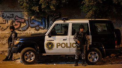 Vehículo policial "POLICE" blanco y negro, flanqueado por dos agentes armados con uniformes de camuflaje y cascos, en una calle nocturna con muro de grafitis árabe. Diciembre 2018, Giza, El Cairo, Egipto. Vehículo policial "POLICE" blanco y negro, flanqueado por dos agentes armados con uniformes de camuflaje y cascos, en una calle nocturna con muro de grafitis árabe. Diciembre 2018, Giza, El Cairo, Egipto.