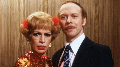 Un fotograma de la serie muestra a una mujer con moños y vestido rojo brillante junto a un hombre con traje oscuro. El fondo es una pared con diseño de madera.