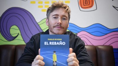 Pablo Rivero muestra su novela "El Rebaño", con portada azul oscuro y título en blanco.  Sentado en un sofá, ante un mural abstracto.
